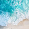 Aerial view of turquoise ocean waves washing onto a sandy beach, with white sea foam and small bird footprints visible on the sand. Beachscape art print available in A1 to A4 sizes.