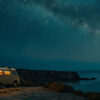 A camper van with interior lights on is parked near a rocky coastline at night under a star-filled sky and the Milky Way. Explorer-themed print available in A1 to A4 sizes.