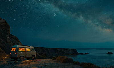 A camper van with interior lights on is parked near a rocky coastline at night under a star-filled sky and the Milky Way. Explorer-themed print available in A1 to A4 sizes.