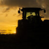 A tractor is silhouetted against a sundown sky with clouds, standing in a field with some grass and fencing visible in the foreground. Nature and landscape poster available in A1 to A4 sizes.