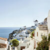 Whitewashed buildings with blue accents on a hillside overlooking the sea, featuring a windmill, under a clear blue sky in Santorini, Greece. Nature and landscape print available in A1 to A4 sizes.
