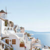 Whitewashed buildings with blue accents on a hillside overlook the sea under a clear sky in Santorini, Greece; a windmill is visible in the background. Nature and landscape print available in A1 to A4 sizes.