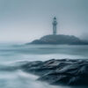 A lighthouse stands on a rocky island in the distance, surrounded by mist and rough, blurred waves in the foreground. Nature and landscape print available in A1 to A4 sizes.