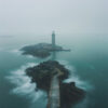A stone pathway leads across rocks to a lighthouse on a small island, surrounded by calm, misty water under an overcast sky. Nature and landscape print available in A1 to A4 sizes.