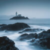 A lighthouse stands on a small rocky island in a misty seascape, with dark rocks and blurred waves in the foreground. Nature and landscape print available in A1 to A4 sizes.