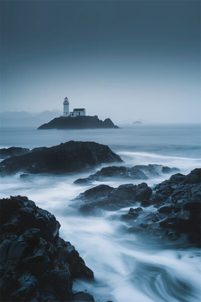 A lighthouse stands on a small rocky island in a misty seascape, with dark rocks and blurred waves in the foreground. Nature and landscape print available in A1 to A4 sizes.