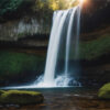 A tall waterfall cascades over a rocky cliff into a pool below, surrounded by moss-covered rocks and lush green vegetation. Sunlight shines through the trees above. Nature and landscape print available in A1 to A4 sizes.