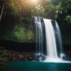 A waterfall flows over a rocky cliff into a turquoise pool, surrounded by dense green vegetation and trees, with sunlight filtering through the foliage. Nature and landscape print available in A1 to A4 sizes.