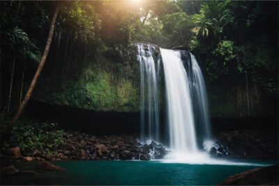 A waterfall flows over a rocky cliff into a turquoise pool, surrounded by dense green vegetation and trees, with sunlight filtering through the foliage. Nature and landscape print available in A1 to A4 sizes.
