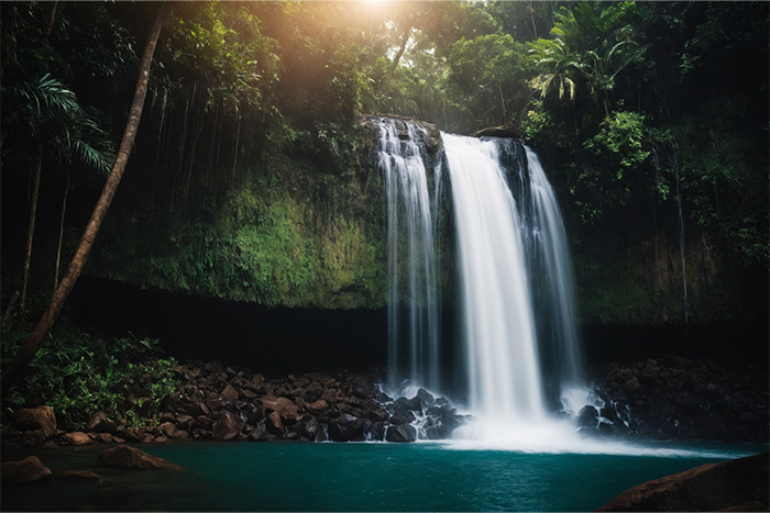 product-image-3-2 A waterfall flows over a rocky cliff into a turquoise pool, surrounded by dense green vegetation and trees, with sunlight filtering through the foliage. Nature and landscape print available in A1 to A4 sizes.