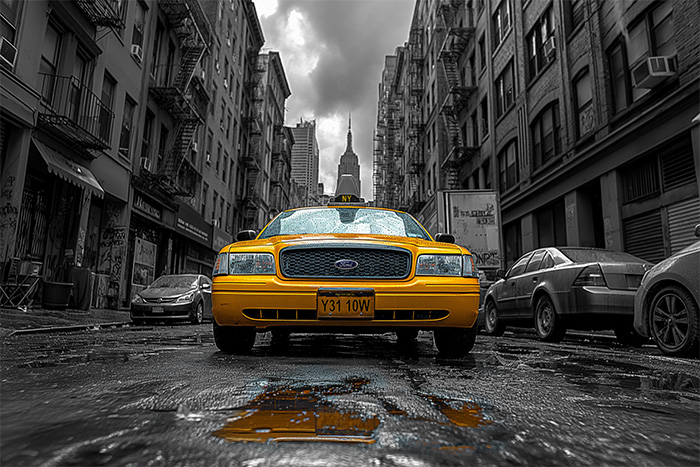 product-image-3-2 A yellow taxi is parked on a wet city street, surrounded by tall, monochrome buildings under a cloudy sky. New York taxi print available in A1 to A4 sizes.