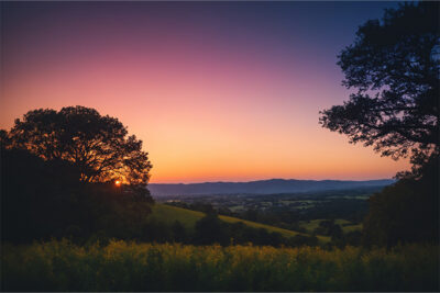Sunset over a rural landscape with rolling hills, trees silhouetted in the foreground, and a gradient sky from orange to purple. Nature and landscape print available in A1 to A4 sizes.
