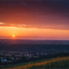 Sunset over a rural landscape with fields, a small village, and distant mountains under a partly cloudy sky. Nature and landscape print available in A1 to A4 sizes.