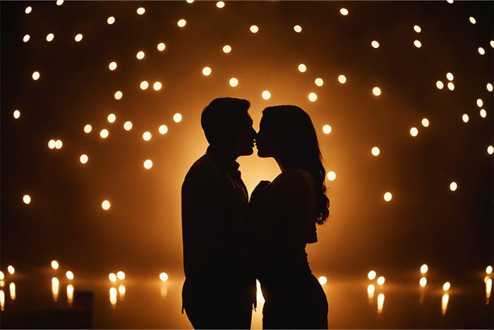 product-image-3-2 Silhouette of a couple about to kiss, standing close together against a backdrop of numerous glowing lights in the dark. Romantic wall art print available in A1 to A4 sizes.