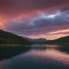 A lake surrounded by hills under a dramatic, colourful sunset sky with clouds reflecting on the water. Nature and landscape print available in A1 to A4 sizes.