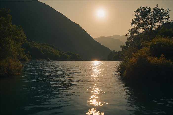 product-image-3-2 A river flows between tree-covered hills at sunset, with sunlight reflecting off the water and mountains visible in the background. Nature and landscape print available in A1 to A4 sizes.