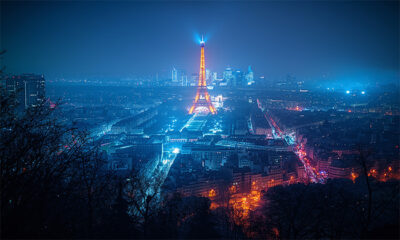 A nighttime cityscape of Paris with the Eiffel Tower illuminated in the centre, surrounded by city lights and buildings under a blue-tinted sky. Wall art print available in A1 to A4 sizes.