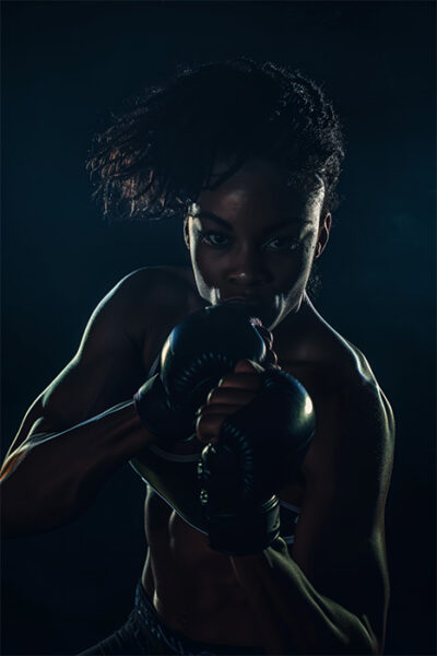 A woman wearing boxing gloves poses in a fighting stance against a dark background, illuminated by dramatic lighting. Poster print available in A1 to A4 sizes.