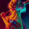 Harley Quinn, with pigtails and face paint, smiling and holding a baseball bat over her shoulder under dramatic lighting. Inspired by the iconic DC Comics character. Fantasy art poster available in A1 to A4 sizes.