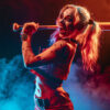 Harley Quinn, in a red and blue costume with pigtails, holds a baseball bat over her shoulder, standing in dramatic lighting as coloured smoke swirls in the background. Fantasy art poster available in A1 to A4 sizes.