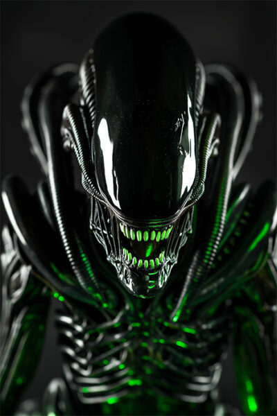 A close-up of a black Alien Xenomorph creature with sharp teeth, shiny surface, and glowing green highlights on its body. Horror poster available in A1 to A4 sizes.