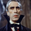 A man in a dark cape and high-collared coat stands indoors, looking serious. His slicked-back hair and vintage attire—a white shirt, red vest, and black cravat—evoke a classic Dracula look. Horror poster available in A1 to A4 sizes.