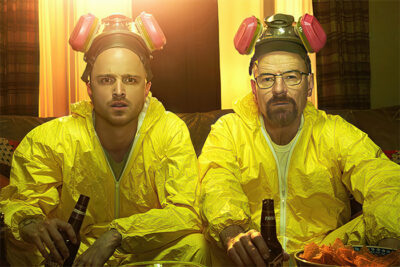 Two men in yellow hazmat suits and respirators sit on a couch, holding beer bottles with a tray of snacks before them, illuminated by warm indoor lighting—a scene from Breaking Bad. Poster print available in A1 to A4 sizes.