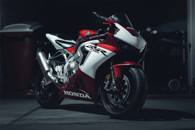 A red and white Honda CBR1000RR sport motorcycle is parked indoors on a concrete floor, with dim lighting highlighting its sleek design and striking details. Wall art print available in A1 to A4 sizes.