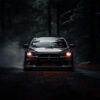 A black Mitsubishi Evo 1 with headlights on is driving along a dark, misty, tree-lined road shrouded in shadows. Wall art print available in A1 to A4 sizes.