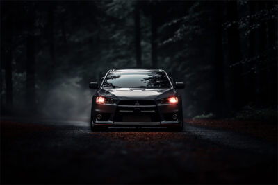 A black Mitsubishi Evo 1 with headlights on is driving along a dark, misty, tree-lined road shrouded in shadows. Wall art print available in A1 to A4 sizes.
