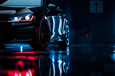 A close-up of a black Volkswagen Golf GTI with illuminated headlights and red accents is reflected on a wet surface in a dimly lit environment. Wall art print available in A1 to A4 sizes.