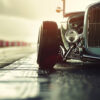 A vintage car with exposed front wheels is parked on a wet racetrack, with the sun shining in the background. Wall art print available in A1 to A4 sizes.