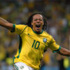 Soccer player Ronaldinho in a yellow Brazil jersey celebrates with arms raised on the field, with a cheering crowd visible in the background. Poster print available in A1 to A4 sizes.