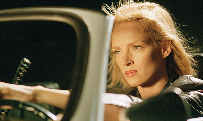 product-image-7-4 Uma Thurman sits in the driver’s seat of a car, looking intently ahead with her hand on the steering wheel, in a scene from the movie Kill Bill. Wall art print available in A1 to A4 sizes.