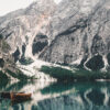 Wooden boats float on the tranquil waters of a Mountain Lake, their reflections mirrored alongside tall, rugged peaks and pine trees in the background. Nature and landscape poster print available in A1 to A4 sizes.