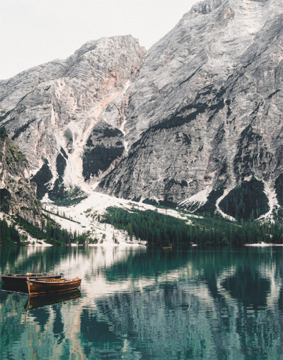 Wooden boats float on the tranquil waters of a Mountain Lake, their reflections mirrored alongside tall, rugged peaks and pine trees in the background. Nature and landscape poster print available in A1 to A4 sizes.