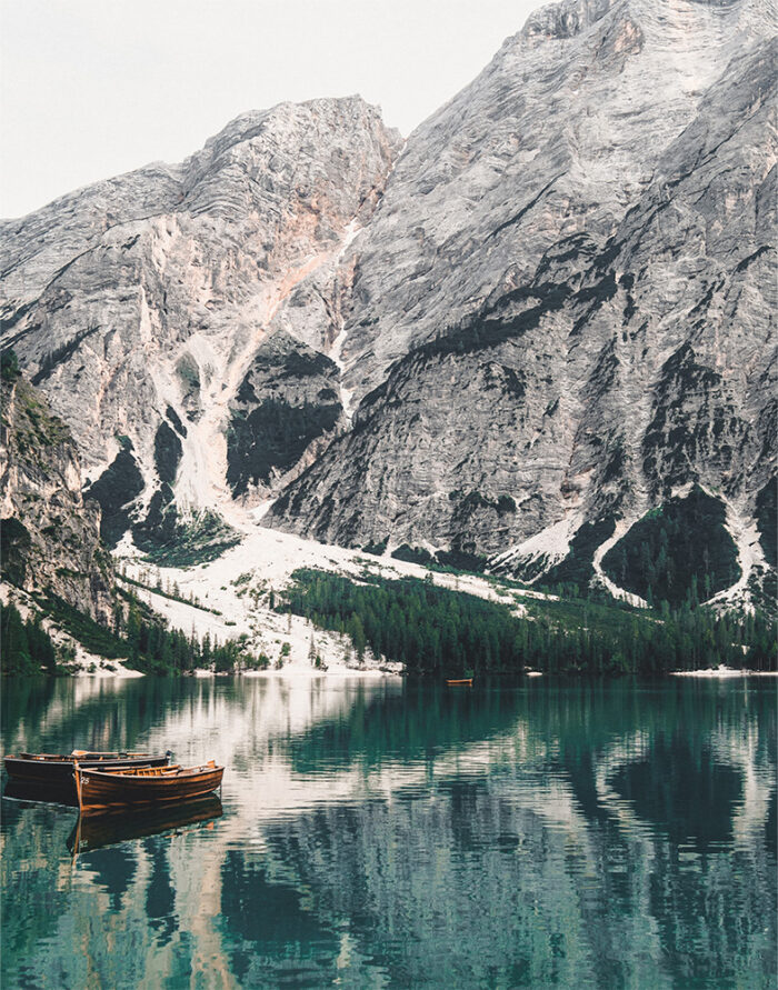 Wooden boats float on the tranquil waters of a Mountain Lake, their reflections mirrored alongside tall, rugged peaks and pine trees in the background. Nature and landscape poster print available in A1 to A4 sizes.