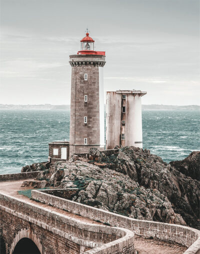 A stone Lighthouse and adjacent building stand on rocky cliffs by the sea, with choppy water and a cloudy sky in the background. Nature and landscape poster print available in A1 to A4 sizes.