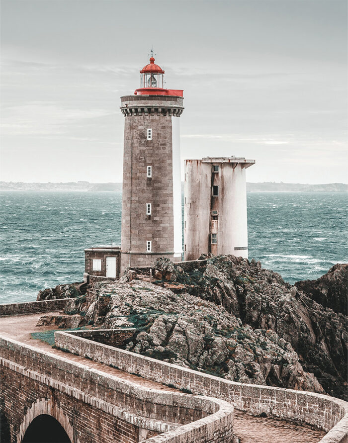 product-image-11-14 A stone Lighthouse and adjacent building stand on rocky cliffs by the sea, with choppy water and a cloudy sky in the background. Nature and landscape poster print available in A1 to A4 sizes.
