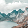 A calm mountain lake bordered by rugged, rocky mountains under a cloudy sky. Small buildings are visible along the shoreline, a serene and picturesque retreat. Nature and landscape poster print available in A1 to A4 sizes.