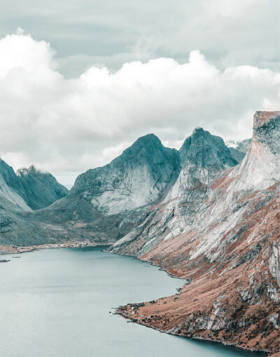 A calm mountain lake bordered by rugged, rocky mountains under a cloudy sky. Small buildings are visible along the shoreline, a serene and picturesque retreat. Nature and landscape poster print available in A1 to A4 sizes.