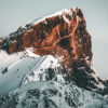 Snow-covered mountain peak with steep, rugged cliffs illuminated by warm sunlight against a pale sky, showcasing the dramatic beauty of a majestic mountain peak. Nature and landscape poster print available in A1 to A4 sizes.