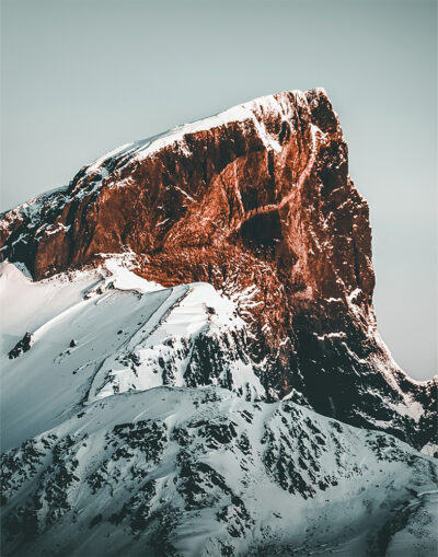 product-image-11-14 Snow-covered mountain peak with steep, rugged cliffs illuminated by warm sunlight against a pale sky, showcasing the dramatic beauty of a majestic mountain peak. Nature and landscape poster print available in A1 to A4 sizes.