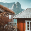 Two red wooden mountain cabins with grass roofs stand by a body of water, with steep rocky mountains rising in the background under a partly cloudy sky. Nature and landscape poster print available in A1 to A4 sizes.