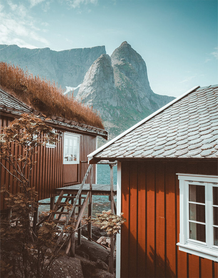 product-image-11-14 Two red wooden mountain cabins with grass roofs stand by a body of water, with steep rocky mountains rising in the background under a partly cloudy sky. Nature and landscape poster print available in A1 to A4 sizes.