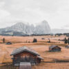 A wooden cabin sits in a grassy field surrounded by Prairie Cabins, scattered huts, rolling hills, and tall mountains rising in the background beneath a cloudy sky. Nature and landscape poster print available in A1 to A4 sizes.