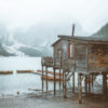 A cozy wooden cabin on stilts stands by the tranquil mountain lake, with rowboats nearby and misty peaks rising behind the trees. Nature and landscape poster print available in A1 to A4 sizes.