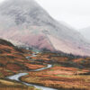 A winding mountain road leads through a hilly landscape with brown grass and scattered rocks, beneath a large, misty mountain under an overcast sky. Nature and landscape poster print available in A1 to A4 sizes.