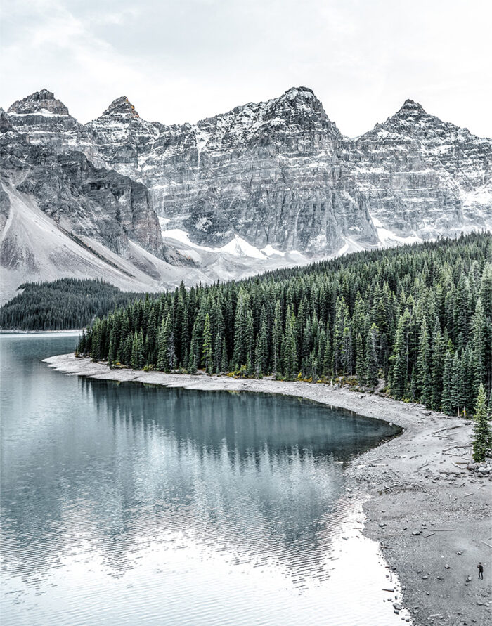 product-image-11-14 A calm Lake is bordered by a dense pine forest, with tall rocky mountains in the background under a cloudy sky. Nature and landscape poster print available in A1 to A4 sizes.