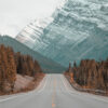 A straight, empty Mountain Road leads through a forest toward a large, snow-capped mountain under a cloudy sky. Nature and landscape poster print available in A1 to A4 sizes.